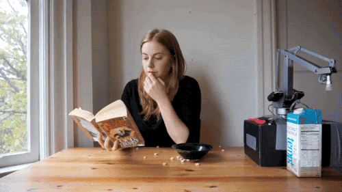 A woman reads a book while a robot tries to pour her cereal and makes a horrible mess.