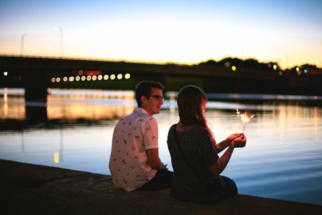 A man and a woman sit by the edge of a river at sundown. The woman holds a sparkler.