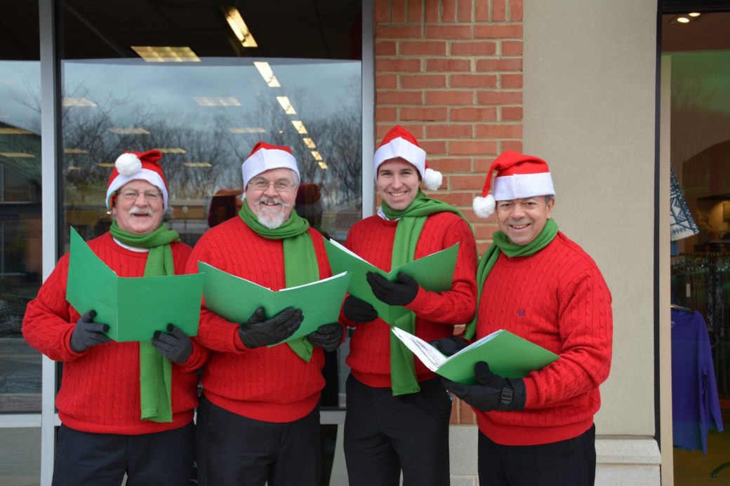 A group of carolers in matching red and green sweatshirts and Santa hats hold their music open in front of a brick wall