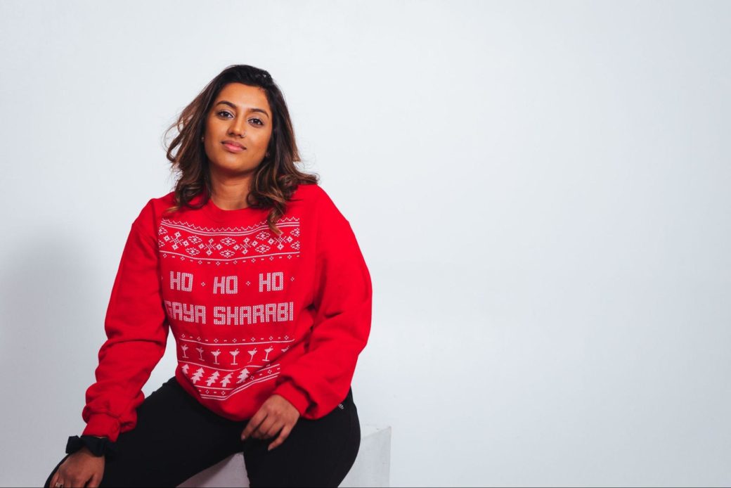 A woman in an ugly sweater sits in front of a white wall