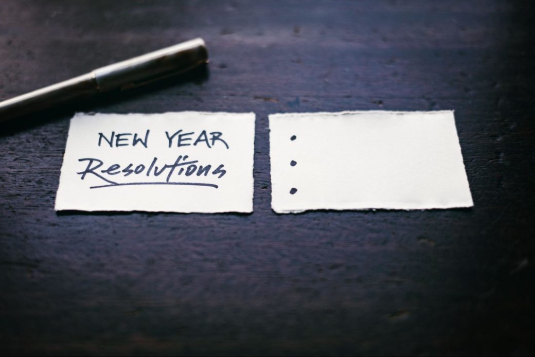 Two slips of paper on a table. One says "New Year Resolutions." The one next to it is blank with bullet points.